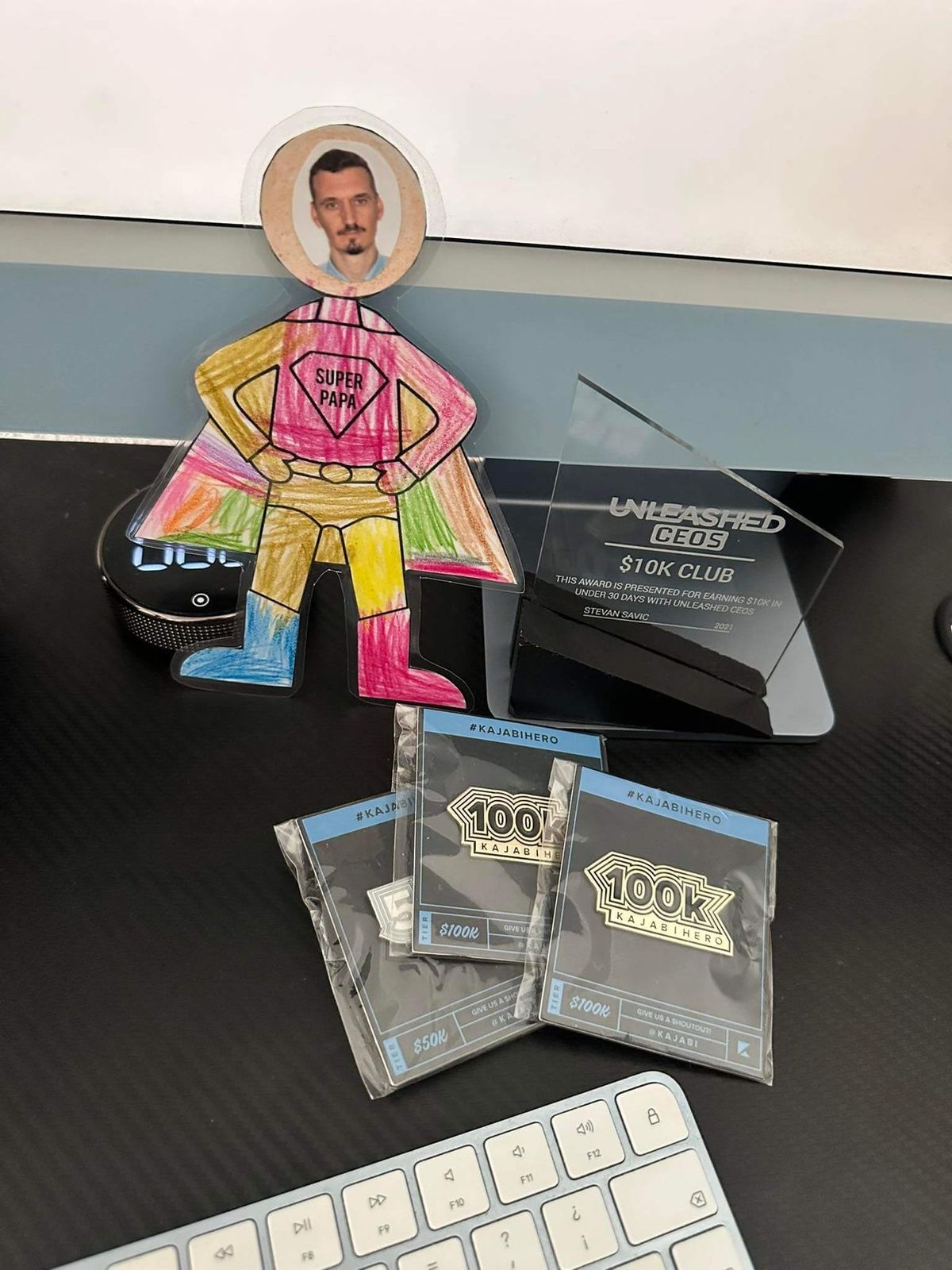 Unleashed CEOs $10K Club award and two Kajabi 100K plaques on my desk, next to my daughter’s Super Papa drawing